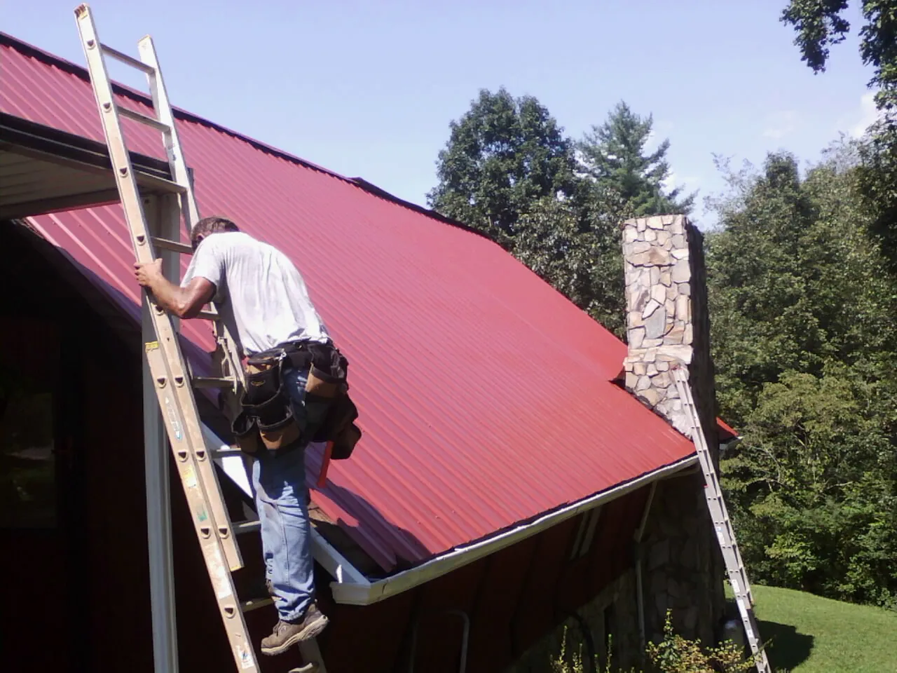 Roofer on a ladder beside a red metal roof and stone chimney — Soffit & Fascia Repair by Roofing Brothers in Carmel
